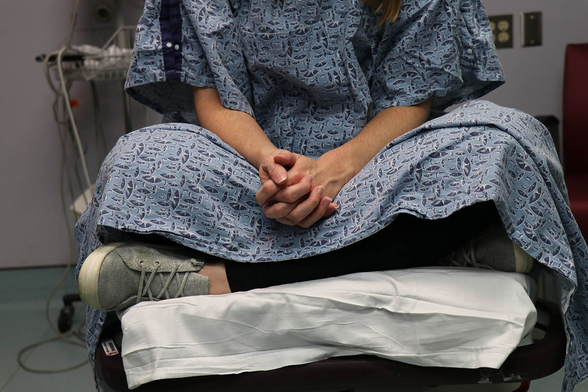 The lower half of an unidentifiable patient is shown wearing a hospital gown and sitting on a table in a hospital room waiting to be examined by a sexual assault nurse examiner (sane)